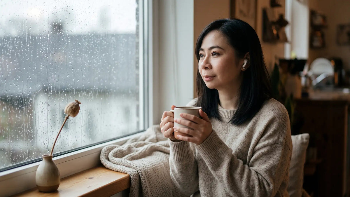 Woman by a rainy window with earbuds and a warm mug, finding quiet from sound sensitivity